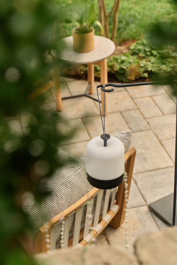 An Acorn Lantern hanging above a teak armchair with a slatted back on a paver patio, with a small side table and plants behind.