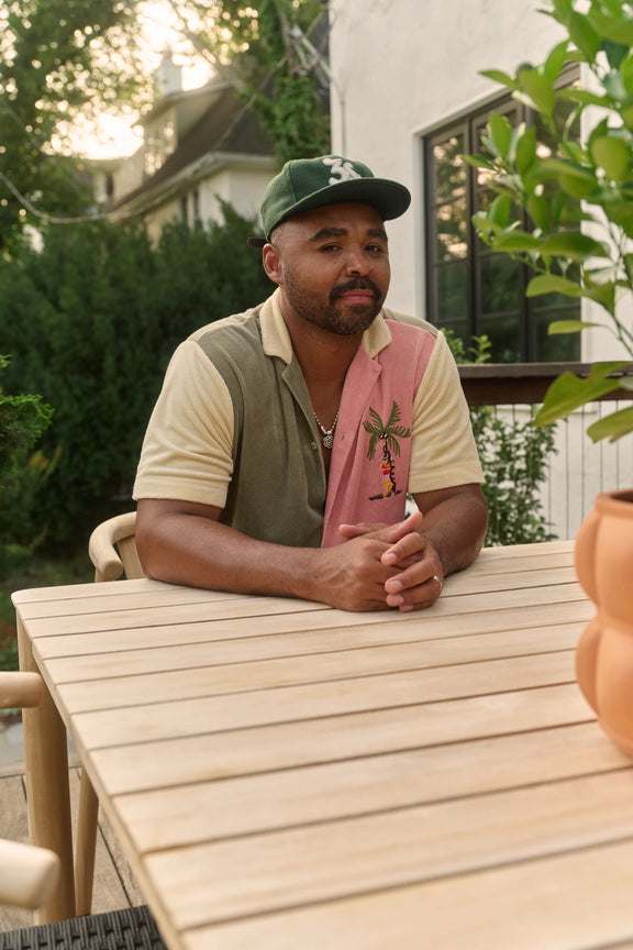 Hilton Carter seated with hands clasped at a teak outdoor dining table, wearing a green cap and a cream-and-pink shirt, with a house and leafy plants behind him.