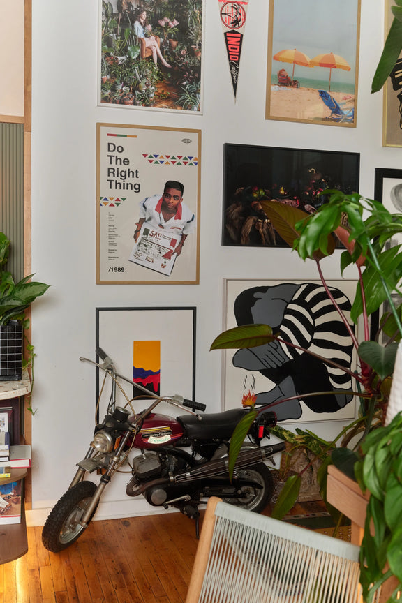 Gallery wall of framed posters and art prints, including a Do the Right Thing poster, beside a motorbike and leafy houseplants.