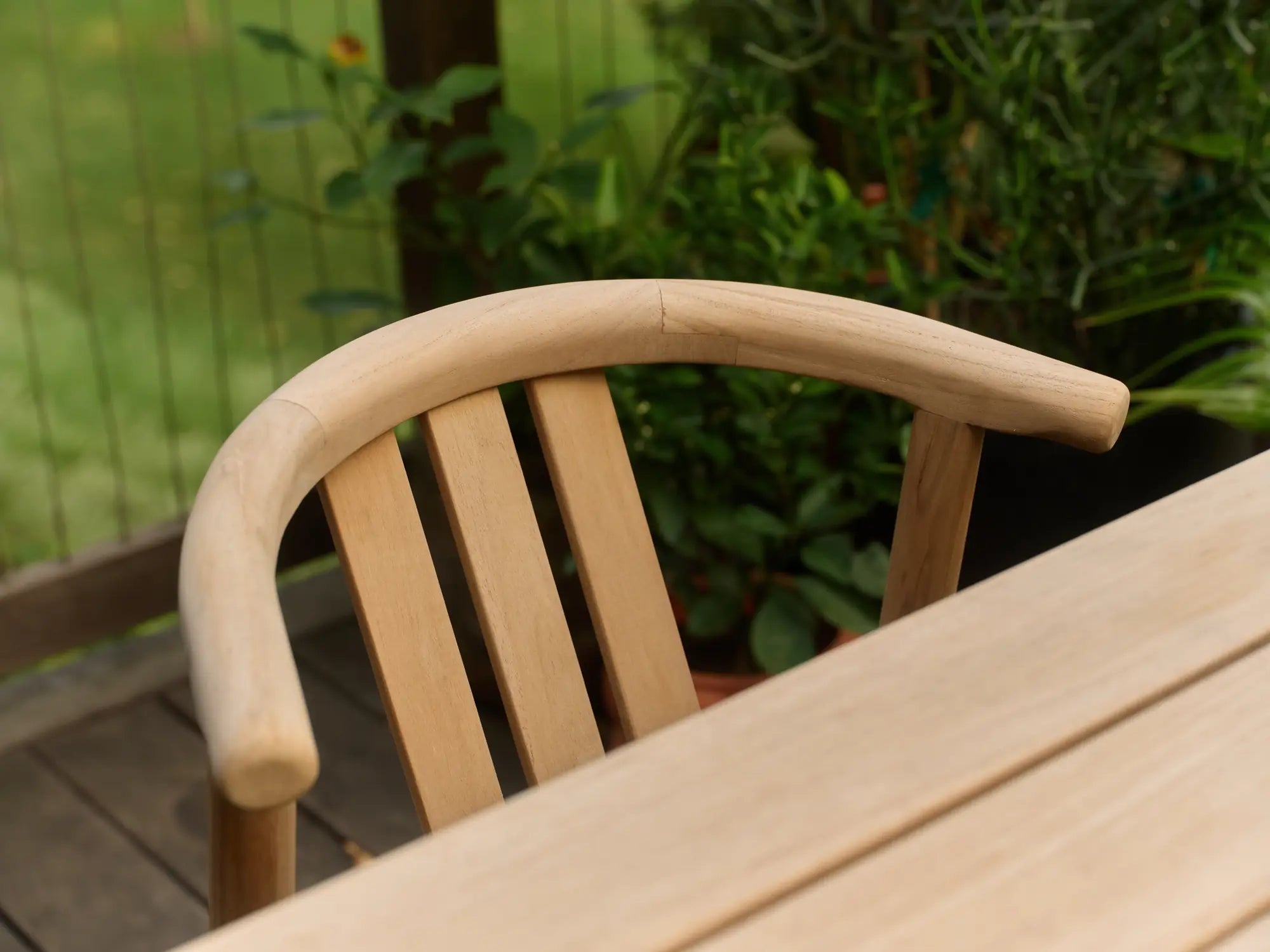 Close-up of a curved teak Dweller chair with a slatted back beside a teak table, with green foliage and a wooden fence blurred in the background.