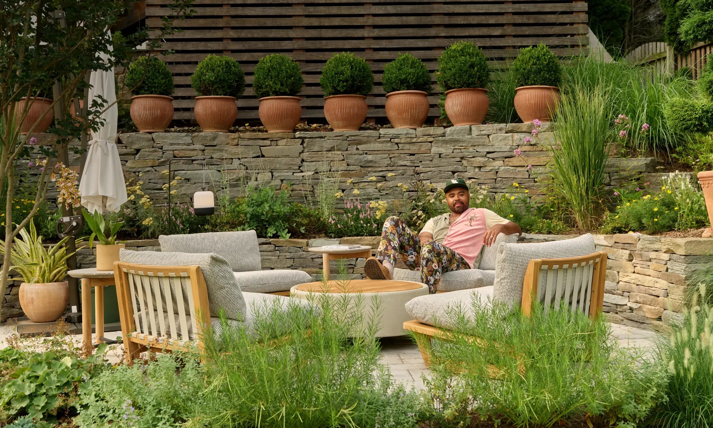 Hilton Carter reclines on a gray-cushioned teak lounge chair in his terraced backyard, with a round concrete fire table, a side table, an Acorn lantern hanging beyond, and terracotta planters along a stone wall