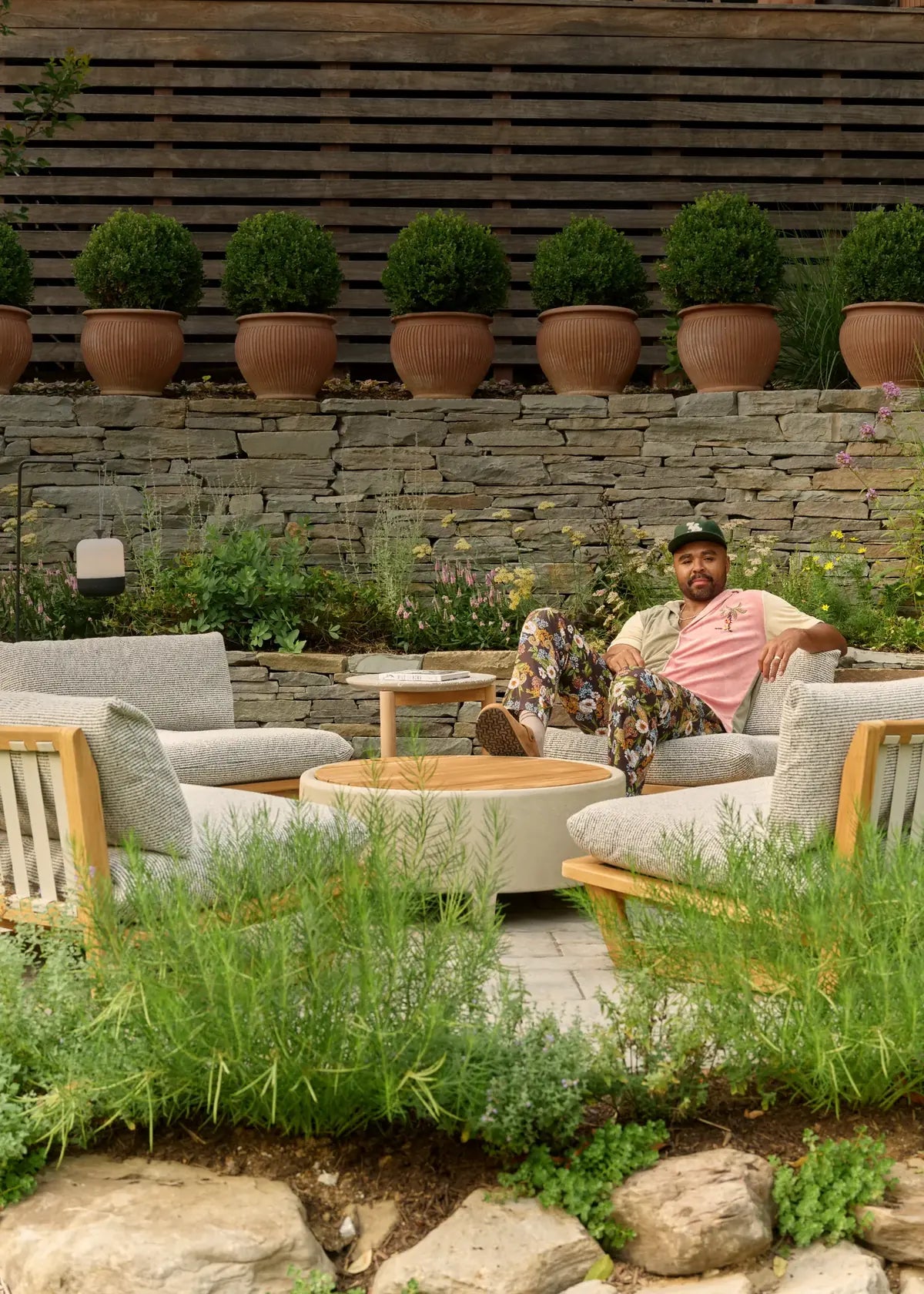 Hilton Carter reclines on a gray-cushioned teak lounge chair in his terraced backyard, with a round concrete fire table, a side table, an Acorn lantern hanging beyond, and terracotta planters along a stone wall