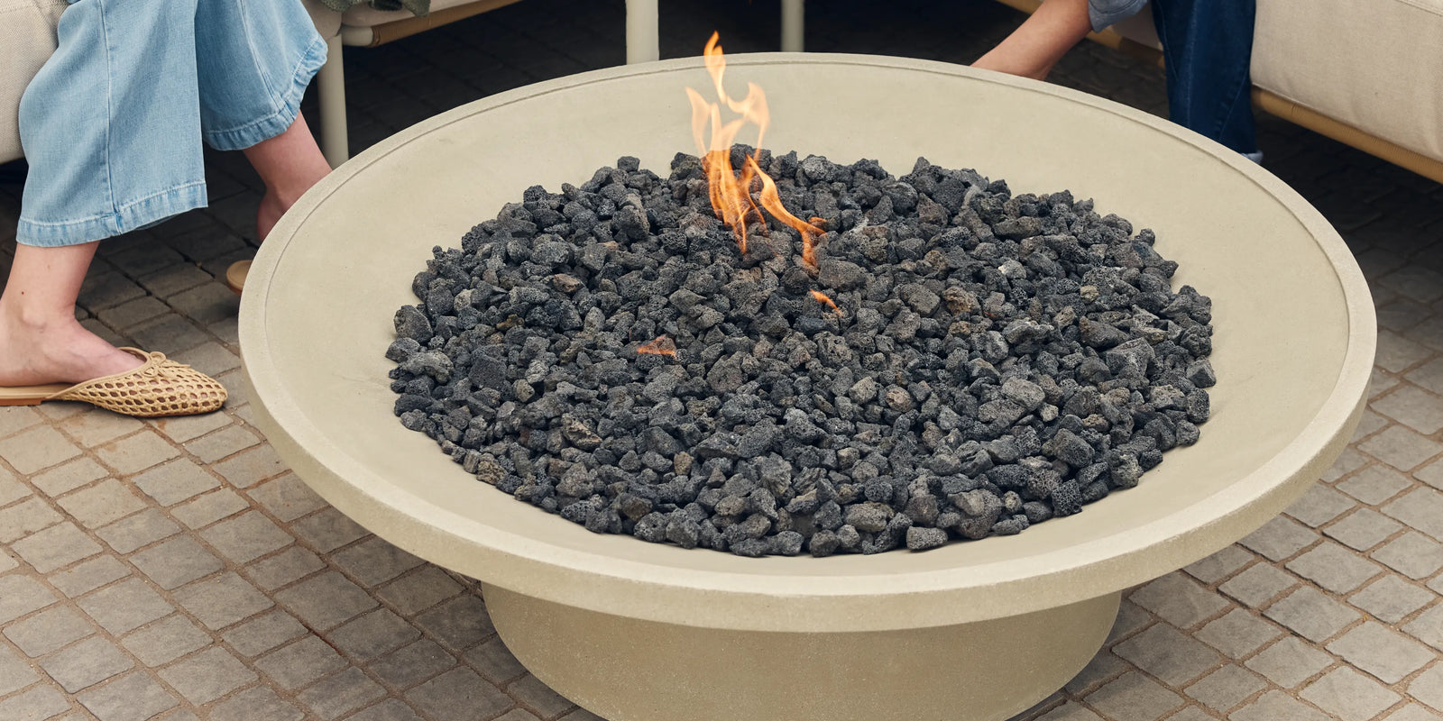 Closeup of Neighbor Brimfire table filled with lava rocks and open flame on a cobblestone patio, Neighbor Camber Sectional visible at edge