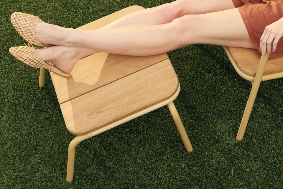 Hopper Ottoman with solid wood construction and curved metal frame, viewed from three-quarter angle on grass