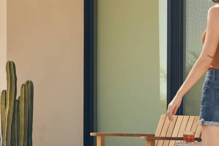 Teak Adirondack chair and ottoman alongside modern house and cactus with a woman partially walking into the frame from the right.