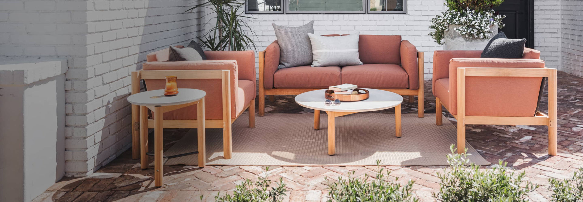 Rust colored cushioned outdoor loveseat made of teak alongside chairs and concrete tables on a bricked patio