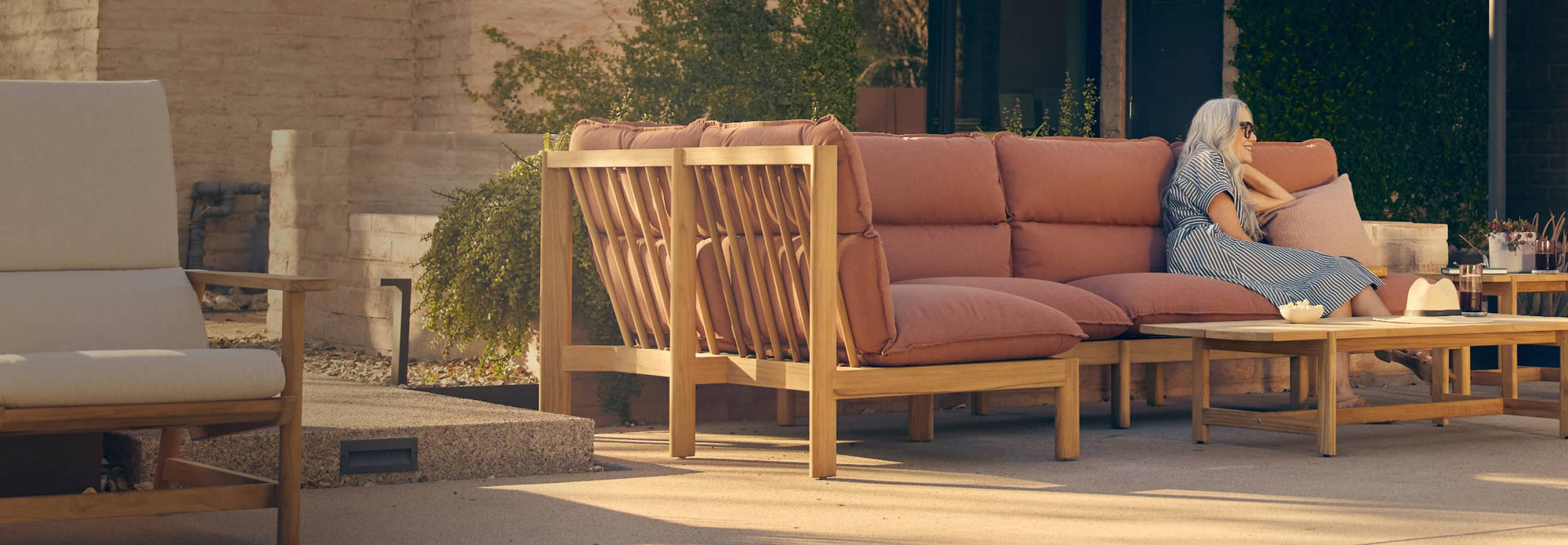 Woman sitting on a comfortable large teak outdoor sectional with rust-colored cushions and teak coffee table