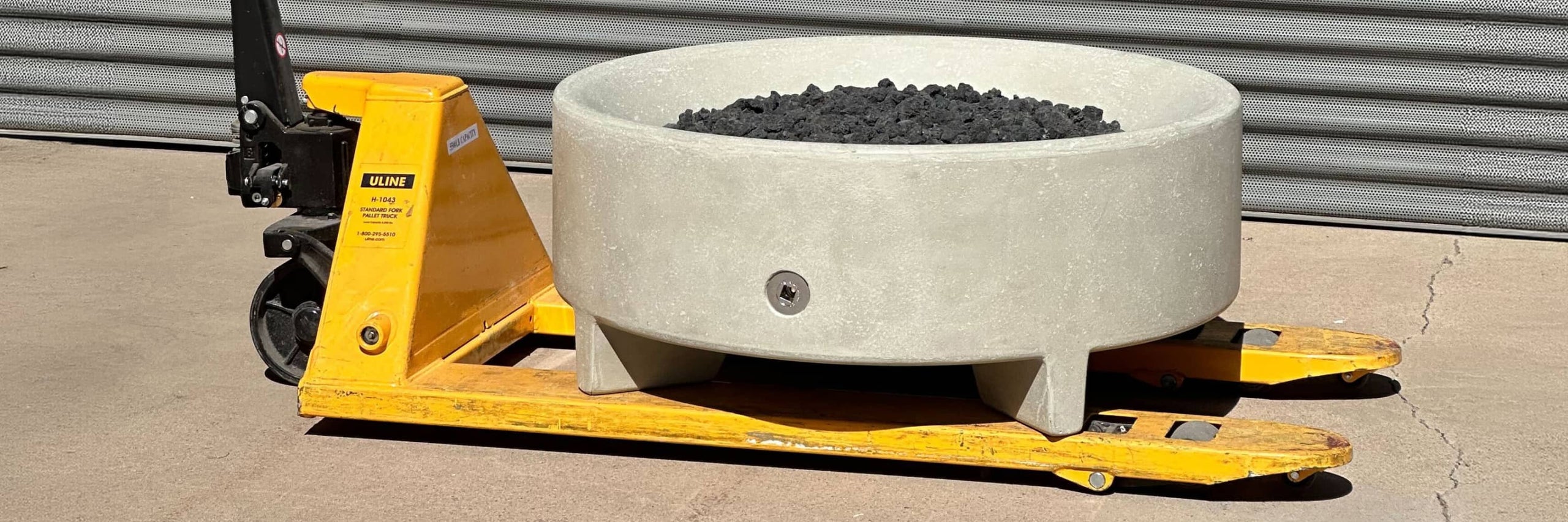 Round concrete fire bowl filled with lava rocks resting on a yellow pallet jack in a warehouse