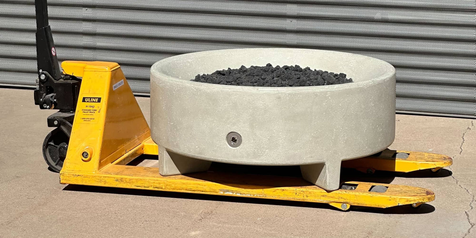 Round concrete fire bowl filled with lava rocks resting on a yellow pallet jack in a warehouse