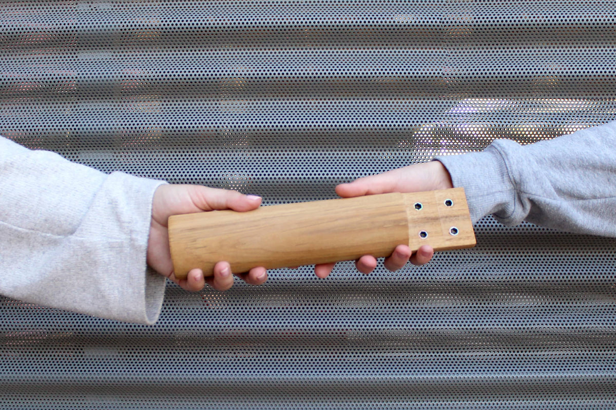 Two people passing a wooden furniture leg between them in front of a perforated metal panel