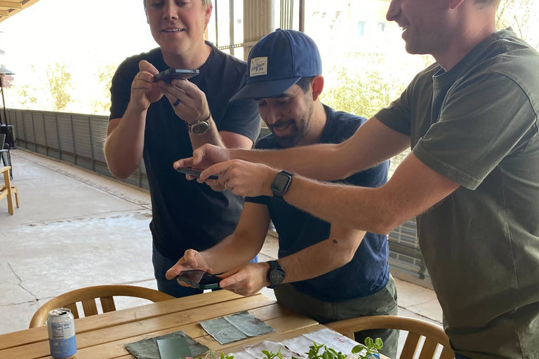 Three people examining material samples and small components together at an outdoor teak table