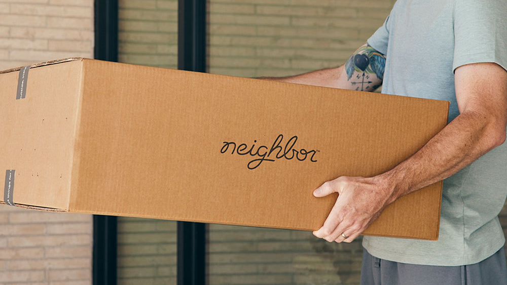 Man carrying a Neighbor-branded cardboard shipping box near a brick building