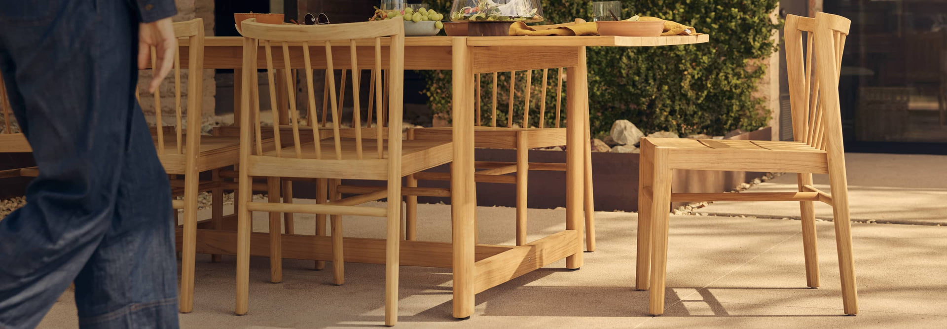 Windsor-styled teak wood dining chair next to a teak dining table on a warm outdoor patio with someone cropped off left edge, walking into frame.