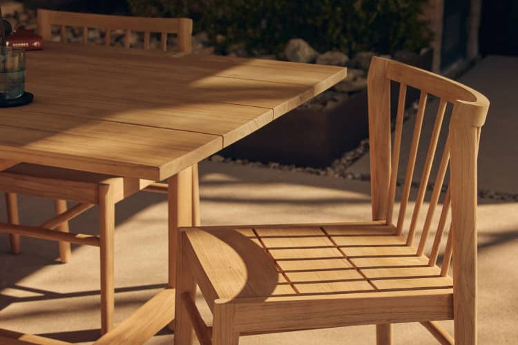 Windsor-styled teak wood dining chair next to a teak dining table on a warm outdoor patio with someone cropped off left edge, walking into frame.